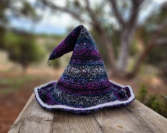 May include: A purple and white striped witch hat with a pointed tip and a wide brim. The hat is made of crocheted yarn and has a white trim around the brim.