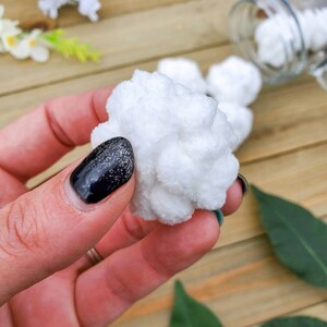 Op de afbeelding: Een close-up van een witte, pluche bal die in een hand met zwarte nagellak wordt vastgehouden. De bal is zacht en lijkt van katoen of garen te zijn gemaakt.