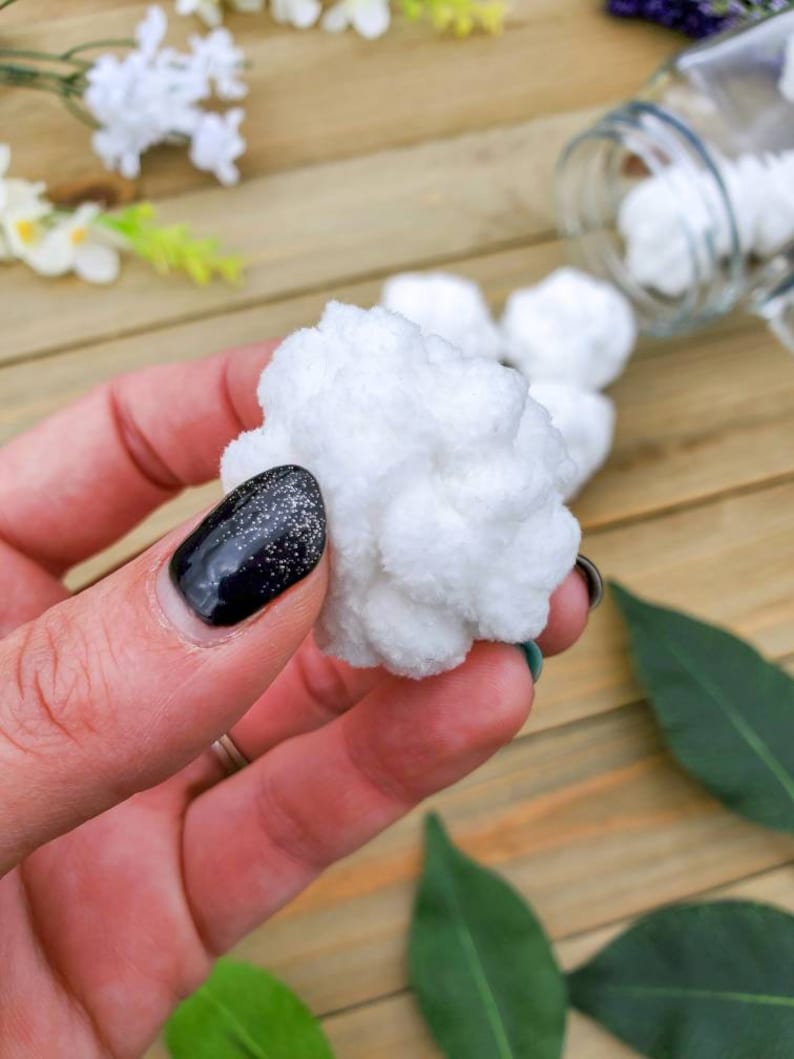 Op de afbeelding: Een close-up van een witte, pluche bal die in een hand met zwarte nagellak wordt vastgehouden. De bal is zacht en lijkt van katoen of garen te zijn gemaakt.