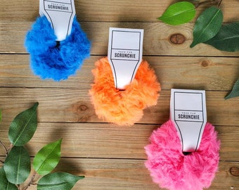 Könnte beinhalten: Drei flauschige Haargummis in Blau, Orange und Pink. Jedes Haargummi hat ein weißes Etikett mit dem Text "Faux Fur Scrunchie" befestigt.