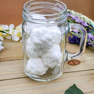 Op de afbeelding: Een glazen mason jar-beker met een handvat gevuld met witte wattenbolletjes. De beker staat op een houten oppervlak met groene bladeren.