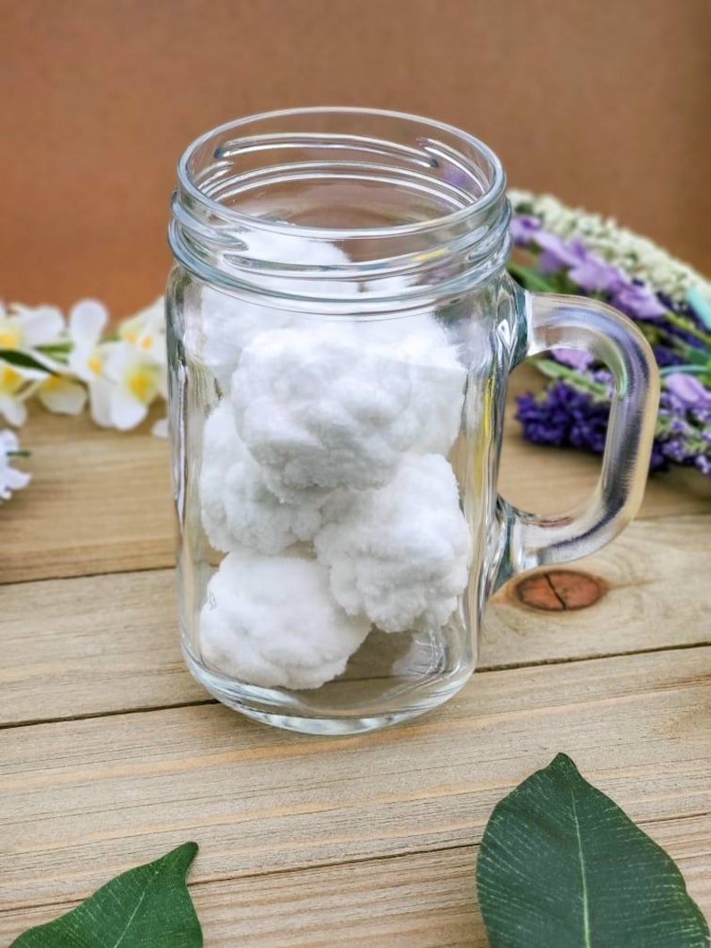 Op de afbeelding: Een glazen mason jar-beker met een handvat gevuld met witte wattenbolletjes. De beker staat op een houten oppervlak met groene bladeren.
