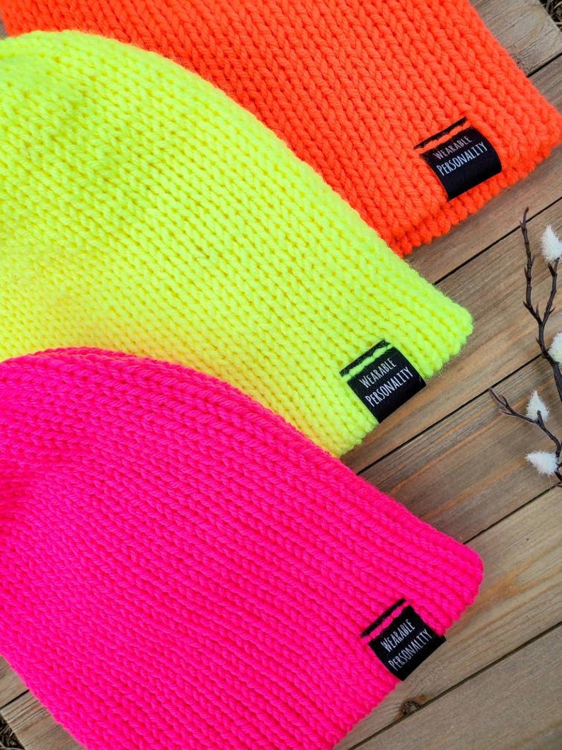 May include: Three knitted beanies in bright neon colors: pink, yellow, and orange. Each beanie has a black label sewn on the side that reads "Wearable Personality."