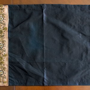 Puede incluir: Un mantel individual rectangular de tela negra con un borde vertical beige en el lado izquierdo. El borde está decorado con intrincados bordados de hilo dorado, pequeñas hojas verdes y motivos florales acentuados con cuentas claras y oscuras.