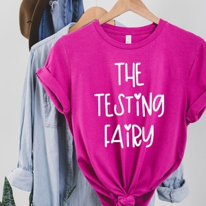 Peut inclure: Un t-shirt rose avec le texte "THE TESTING FAIRY" en blanc avec de petits cœurs de chaque côté des mots "TESTING" et "FAIRY".