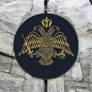 Op de afbeelding: Een ronde, marineblauwe patch met een goud geborduurd dubbelkoppige adelaar, kroon, zwaard en bol ontwerp. De patch is geplaatst tegen een achtergrond van getextureerde grijze steen.