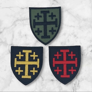 May include: Three embroidered patches in a shield shape. One is olive green with a black cross design. The other two are navy blue with a gold and red cross design. The patches are on a white marble surface.