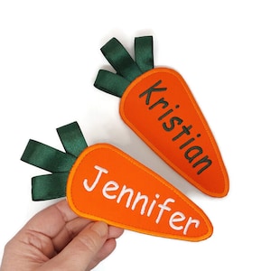 Op de afbeelding: Twee vilten carotvormige ornamenten met groene vilten toppen. De wortels zijn oranje en hebben de namen "Jennifer" en "Kristian" geborduurd in witte draad.
