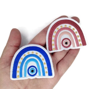 Puede incluir: Dos parches bordados con forma de arcoíris con un ojo azul mal de ojo en el centro. El arcoíris está hecho de tela azul, rosa y roja con estrellas doradas alrededor de los bordes.