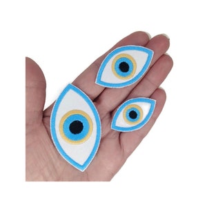 May include: Three blue evil eye patches in different sizes. The patches are white with a blue outline and a blue eye with a yellow ring and a black pupil.
