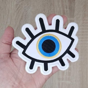 Può includere: Toppa ricamata bianca a forma di occhio con un'iride blu e oro. L'occhio è contornato di nero e ha uno sfondo bianco.
