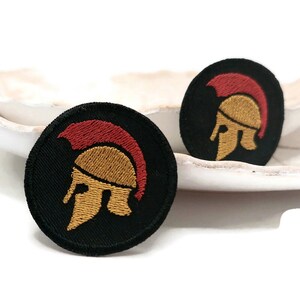 Könnte beinhalten: Zwei schwarze, gestickte Bügelbilder mit einem goldenen Spartanerhelm-Design. Der Helm hat eine rote Feder.