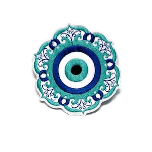 May include: A white embroidered patch with a blue and turquoise evil eye design. The evil eye is surrounded by a decorative floral pattern.