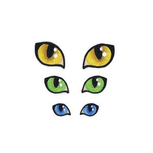 May include: A set of embroidered patches depicting cat eyes in three sizes and colours. The top pair are yellow, the middle pair are green, and the bottom pair are blue. Each eye has a black outline and a black pupil.