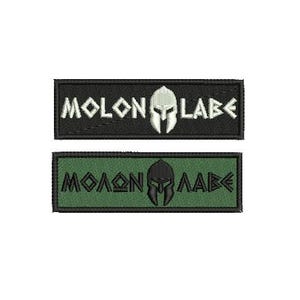 Könnte beinhalten: Zwei gestickte Aufnäher. Der obere Aufnäher ist schwarz mit weißem Text und einem weißen spartanischen Helm. Der Text lautet "MOLON LABE". Der untere Aufnäher ist grün mit schwarzem Text und einem schwarzen spartanischen Helm. Der Text lautet "MOLON LABE".