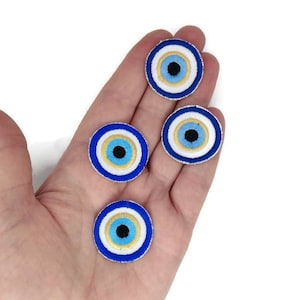 Peut inclure: Quatre écussons brodés représentant l'œil protecteur. Chaque écusson a un anneau extérieur bleu, un anneau intérieur blanc, un anneau doré, un anneau bleu clair et un centre noir. Les écussons sont ronds.