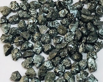 May include: A collection of small, dark grey, rough-cut diamonds. The diamonds are scattered on a white surface.