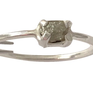 May include: A silver ring featuring a raw, uncut diamond. The diamond is a light gray color and is held in place by four prongs. The band is smooth and simple, creating a minimalist design.