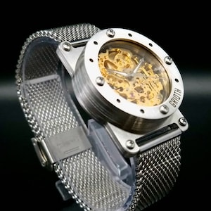 May include: A silver watch with a mesh strap and a clear face revealing the gold-toned inner workings. The watch face has a black rim with small silver dots and the word "GROOTH" is printed on the side.