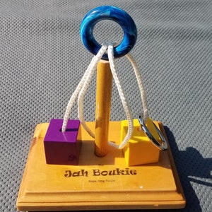 May include: A wooden puzzle called "The Jah Boukie Rope Ring Puzzle". The puzzle has a wooden base, a wooden post, and three colored blocks. The blocks are purple, yellow, and blue. The puzzle also has a blue ring and white rope.