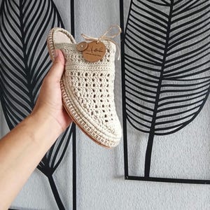 Może przedstawiać: Para beżowych, szydełkowych kapci z brązową skórzaną metką z napisem "Yola".