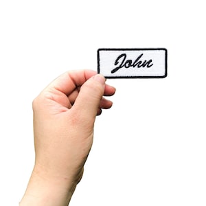 May include: A white rectangular patch with a black embroidered name "John" in a cursive font.