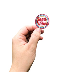 May include: A round embroidered patch with a white background and red, yellow, and blue concentric circles. The text "Good Vibes" is written in red cursive lettering.