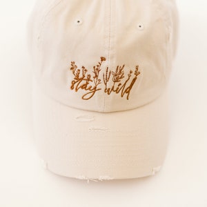 May include: A beige baseball cap with distressed edges and brown embroidery that reads "stay wild" with a floral design.