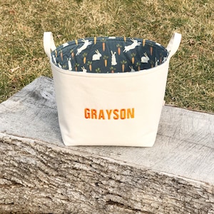 May include: A white canvas storage bin with a blue and white bunny and carrot print lining. The bin has a personalized name, "GRAYSON", embroidered on the front in orange.