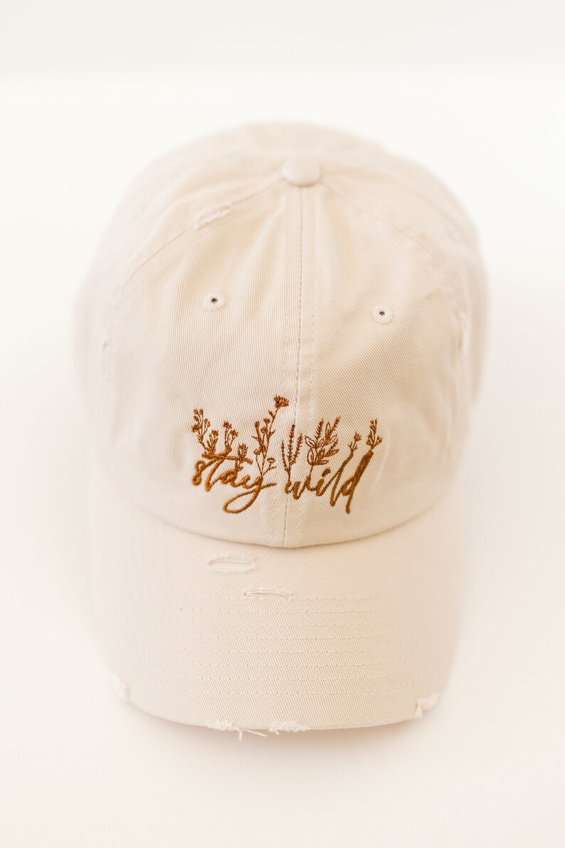 May include: A beige baseball cap with distressed edges and brown embroidery that reads "stay wild" with a floral design.