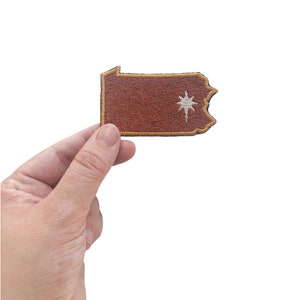 May include: A brown embroidered patch in the shape of the state of Pennsylvania, with a gold border and a white starburst design. The patch is held in a hand against a white background.