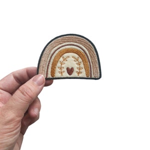 May include: A beige, brown, and orange embroidered rainbow patch with a small red heart in the center and green leaves on either side of the heart.