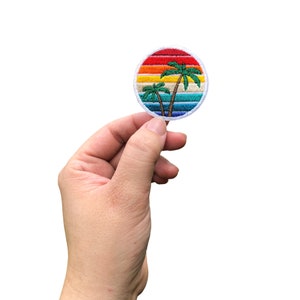 May include: A round embroidered patch with a sunset and palm trees. The sunset is a rainbow of colors, including red, orange, yellow, green, blue, and purple. The palm trees are green and have brown trunks.
