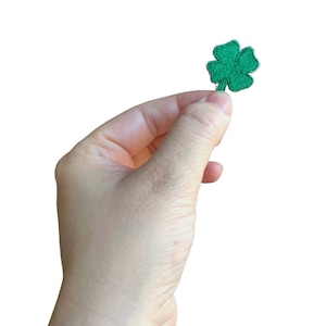 May include: A green four-leaf clover patch, held in a hand, against a white background.