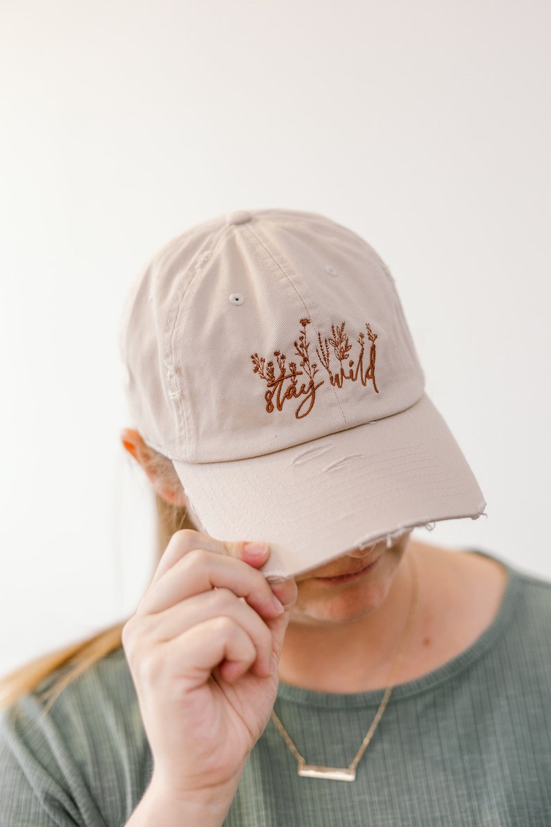 May include: A beige baseball cap with a distressed brim. The cap has brown embroidery that reads "stay wild" with a floral design.