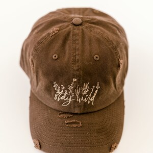 May include: A brown distressed baseball cap with the text "stay wild" embroidered in a light brown color. The text is surrounded by a small floral design.