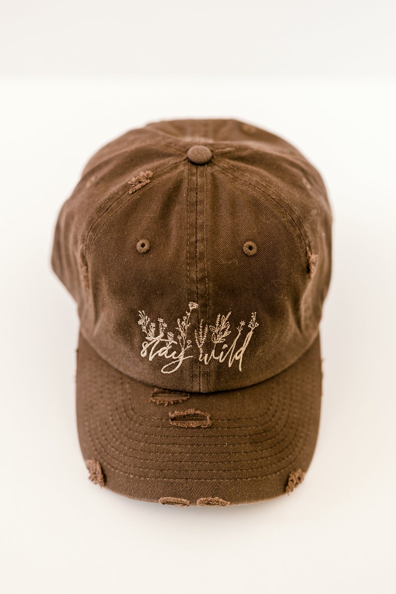 May include: A brown distressed baseball cap with the text "stay wild" embroidered in a light brown color. The text is surrounded by a small floral design.
