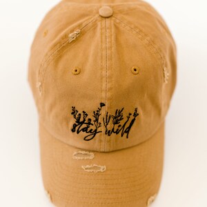 May include: A tan distressed baseball cap with black embroidery that reads "stay wild" with a floral design.