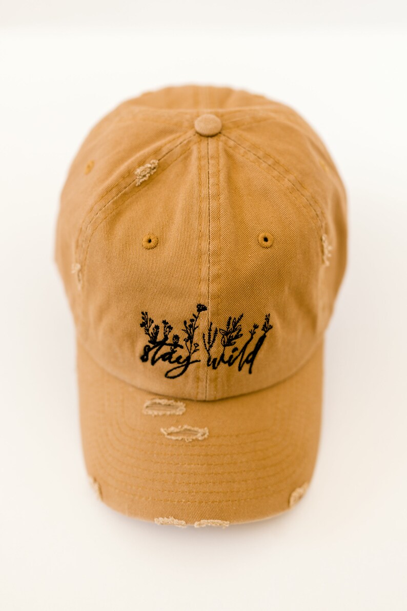 May include: A tan distressed baseball cap with black embroidery that reads "stay wild" with a floral design.