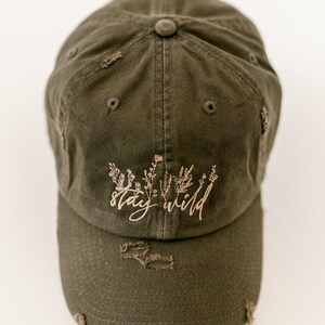 May include: A green distressed baseball cap with the text "stay wild" embroidered in brown on the front. The cap has a brown floral design embroidered above the text.