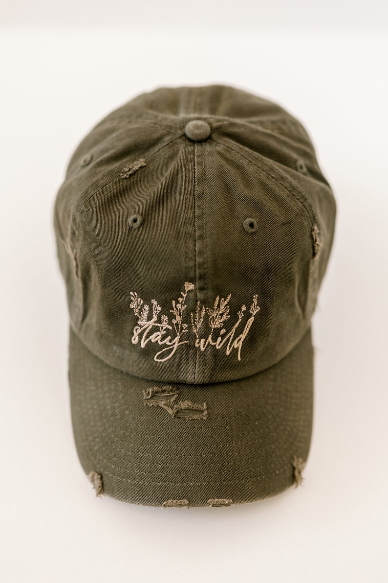 May include: A green distressed baseball cap with the text "stay wild" embroidered in brown on the front. The cap has a brown floral design embroidered above the text.