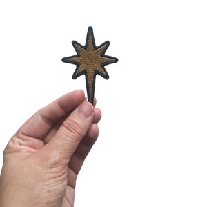 May include: A gold star-shaped patch with a black outline. The patch is held in a hand.