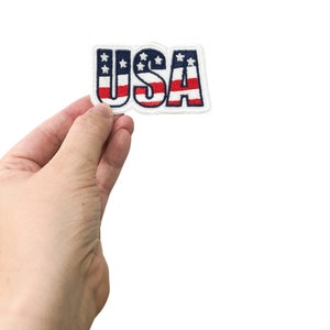 May include: A hand holding a white patch with the word "USA" in red, white, and blue letters. The letters are outlined in blue and have a pattern of red and white stripes and white stars.