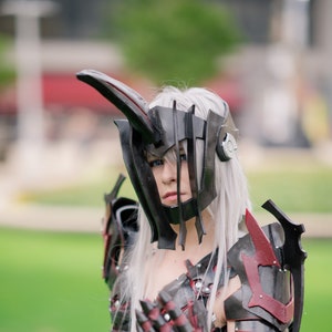 May include: A person in a cosplay outfit featuring a black and red armored chest piece, gauntlets, and helmet with a horn-like extension. The costume includes a black cape and white hair, set against a green background.