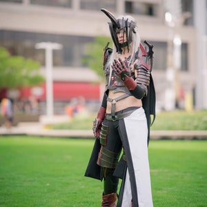 Puede incluir: Una persona con un detallado disfraz de cosplay está de pie sobre hierba verde. El disfraz presenta un casco con cuerno, armadura y una falda negra y blanca. La combinación de colores incluye detalles en negro, rojo y plata, con botas marrones.