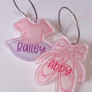 May include: Two pink acrylic keychains with silver cable rings. One keychain features a pink ballerina dress with the name "Bailey" and the other keychain features a pair of pink ballet shoes with a pink bow and the name "Abby".
