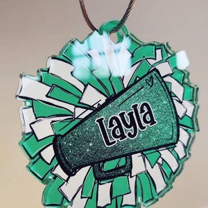 May include: Green and white cheerleading pom-pom ornament with a green glitter megaphone that says "Layla" on it.