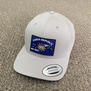 May include: A gray and white mesh trucker hat with a blue and gold patch that says "Conch Republic 1829 Key West". The hat also has a white label that says "The Authentic Yupoong Snapback Since 1974".