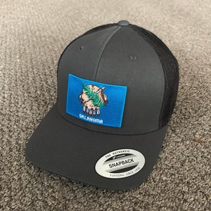 May include: A black and gray trucker hat with a blue and white Oklahoma state flag patch on the front. The hat has a black mesh back and a black snap closure. The hat also has a white oval sticker with the text "The Authentic Yupoong Snapback Yupoong Since 1974".