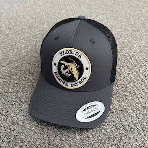 Puede incluir: Una gorra de camionero de malla gris y negra con un parche bordado en dorado que dice "Florida Marine Patrol" con un ancla negra y un contorno dorado del estado de Florida.